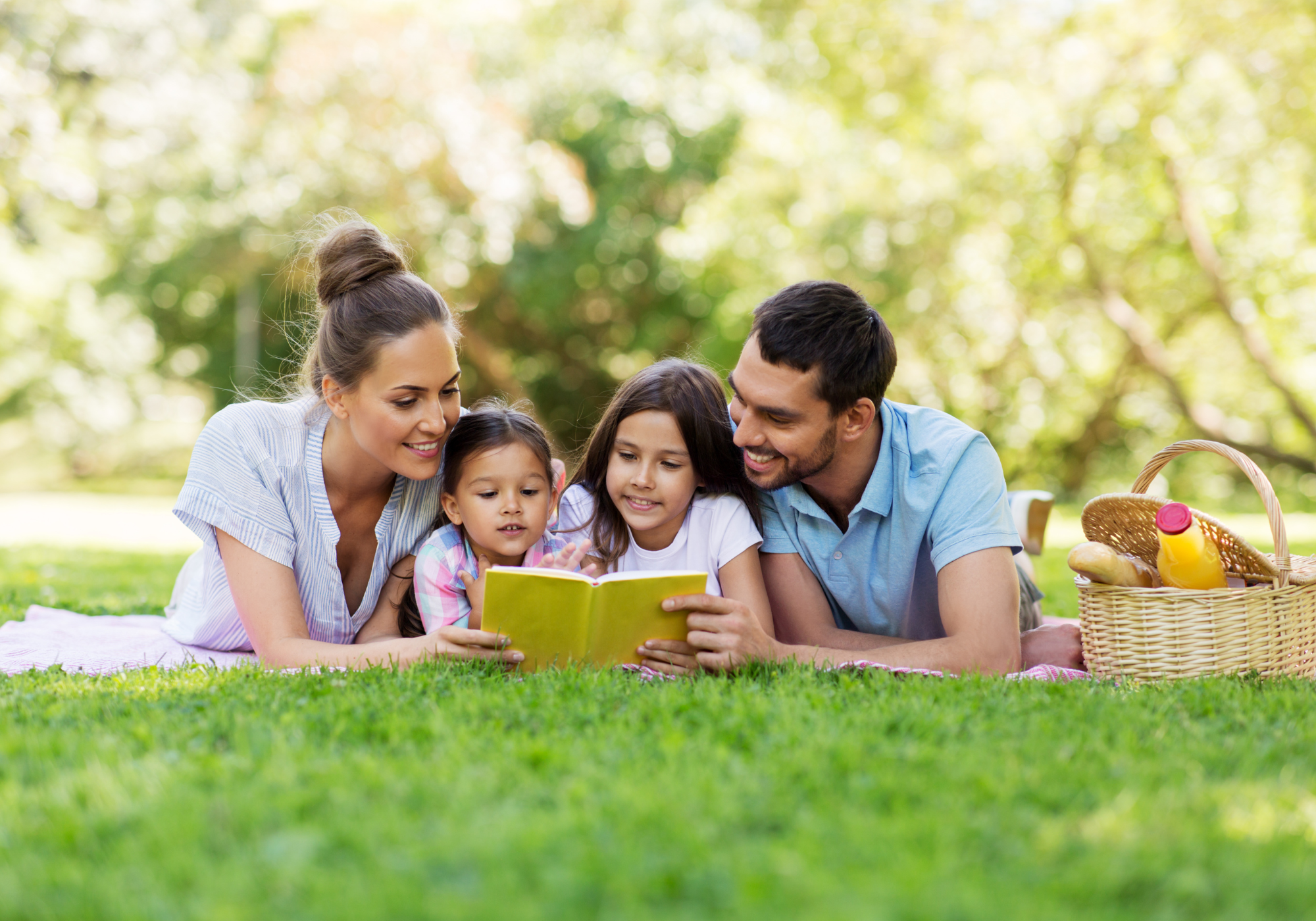 A family enjoying a sunny day at the park, with some members having a picnic on a blanket and others reading a book under a tree.